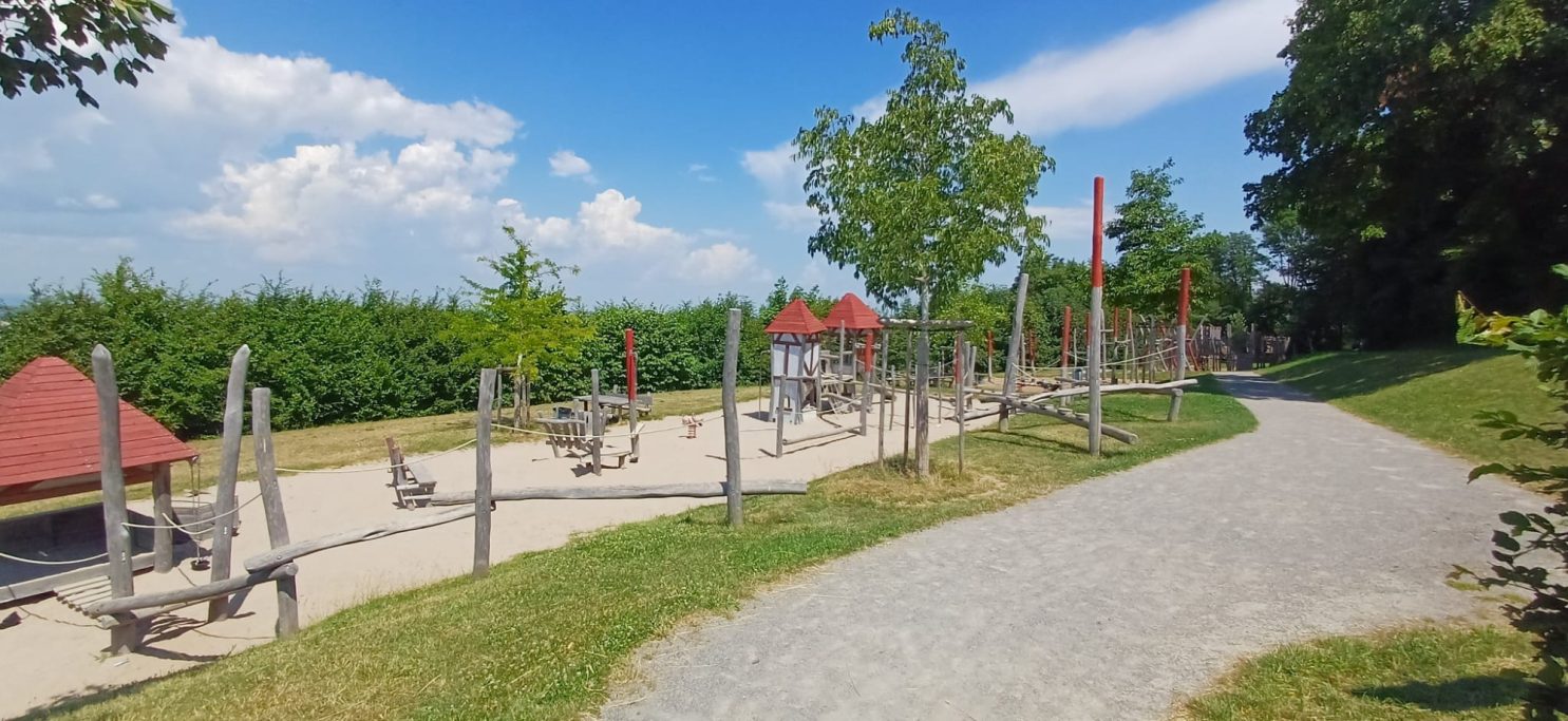 Spielplatz Spielplatz mit Geräten, Sandfläche und Bäumen unter blauem Himmel.