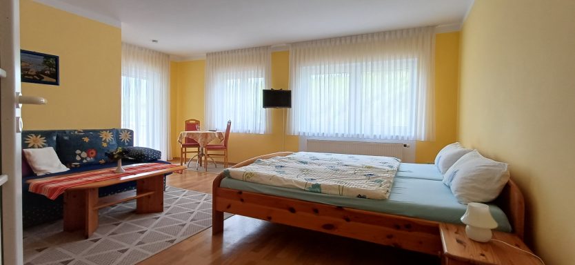 Schlafzimmer 1 Helles Schlafzimmer mit zwei Betten, Sofa und Tisch, umlaufend große Fenster.
